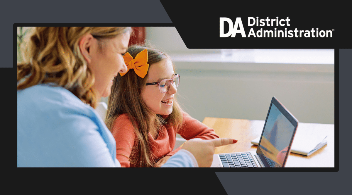 Female teacher pointing at laptop screen with young female student in glasses laughing. District Administration logo on top right.