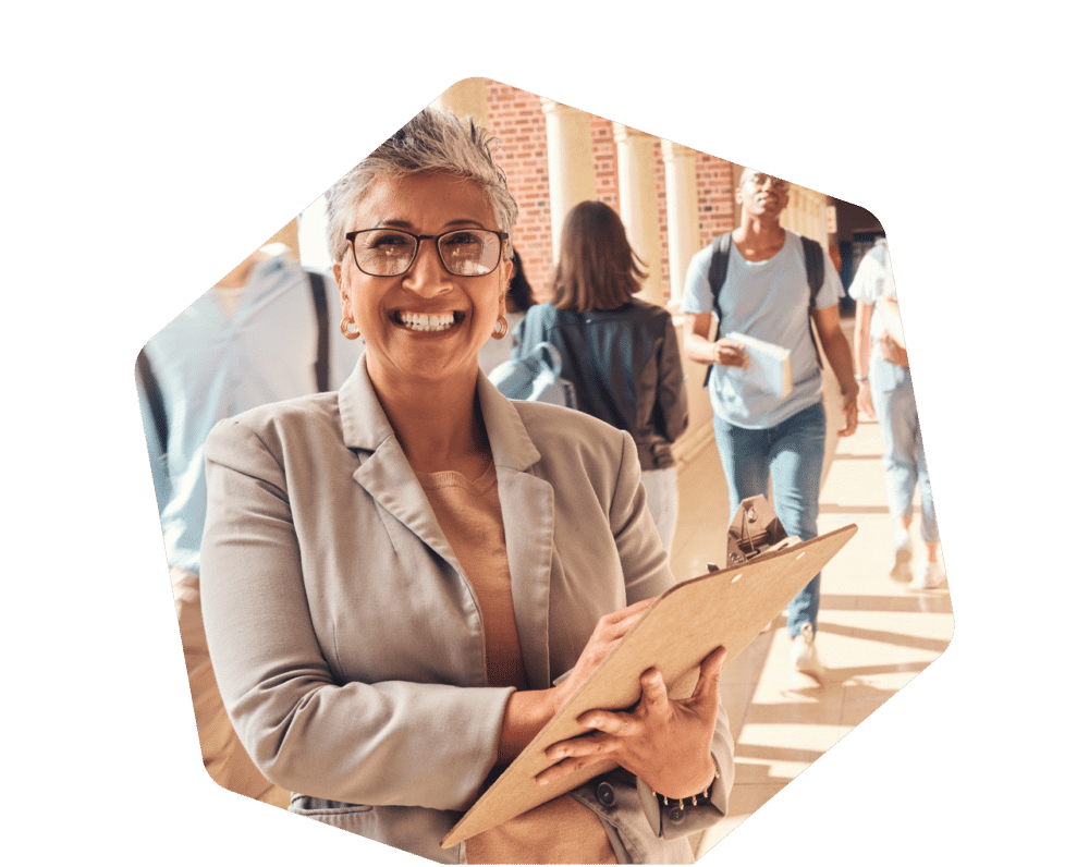 Smiling district leader holding a clipboard, standing in a busy school hallway with students walking in the background.